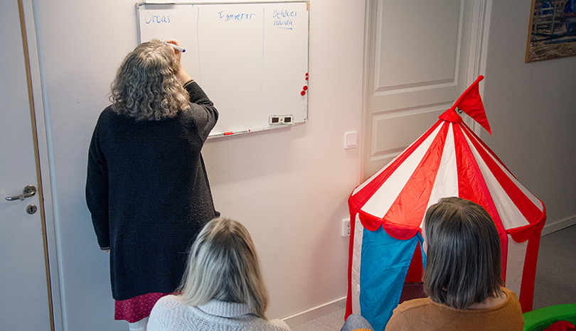 Kvinna skriver på whiteboardtavla. Två andra kvinnor sitter och tittar på.